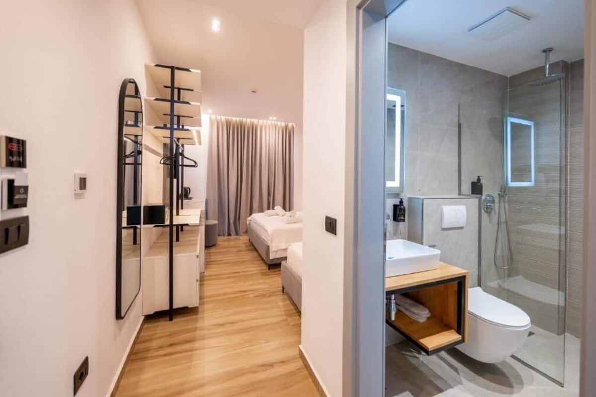 A modern hotel room with a wooden floor, a bed with white linens, a glass shower, and a small wooden shelf under the sink.
