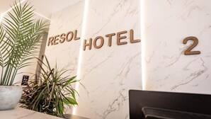 Vastaanottotiski, jonka takana on marmoriseinä, siinä on kasvi ja teksti: ”RESOL HOTEL 2”.