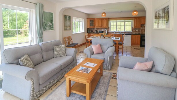 A spacious living area with a sofa set, a coffee table, and a dining table with chairs. There is a kitchen in the background with wooden cabinets and a window. - THE SKATING ROCK, pet friendly in Kenmare, County Kerry (Bonane)