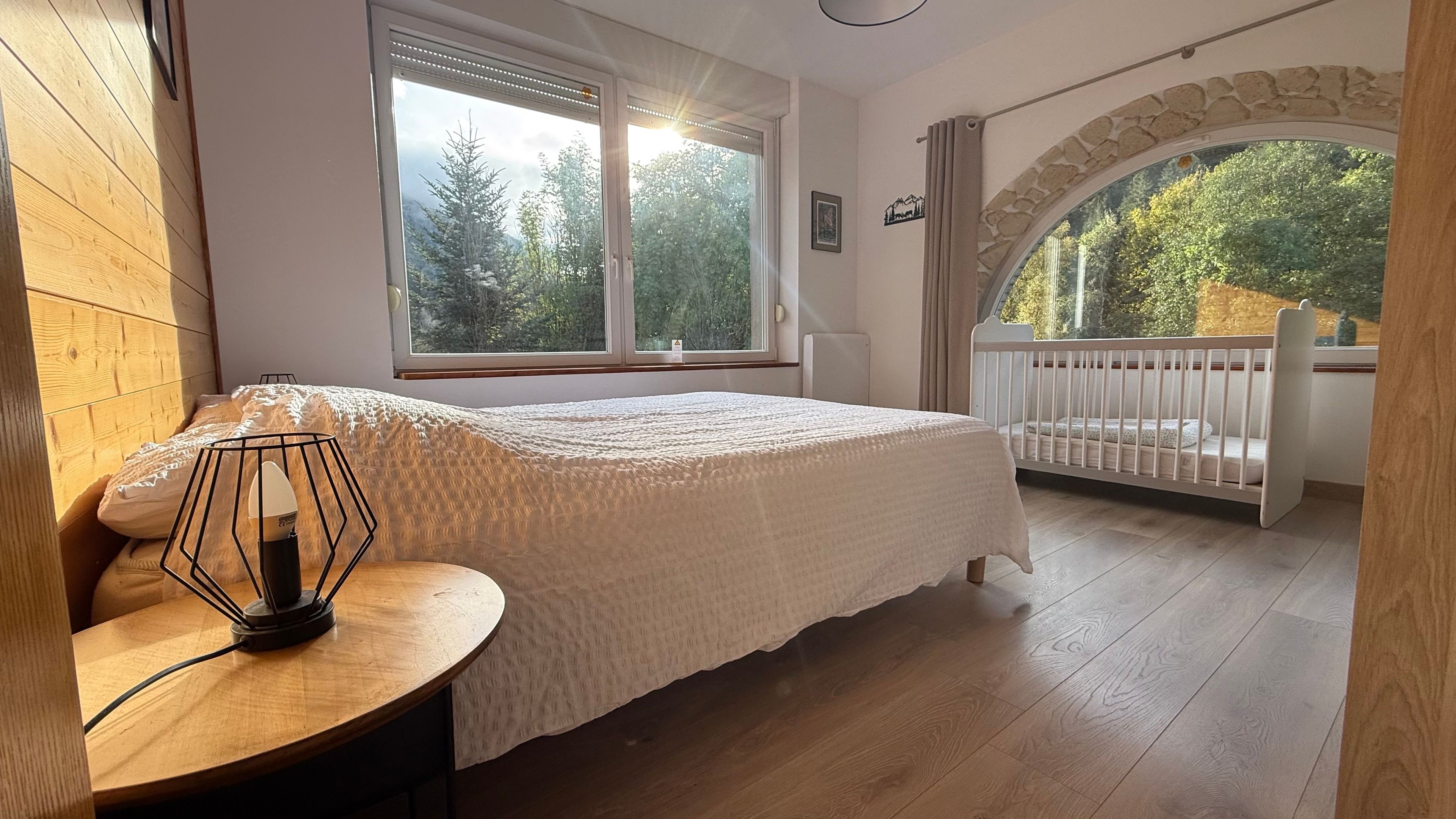 A bedroom with a large window, a crib, and a wooden floor.