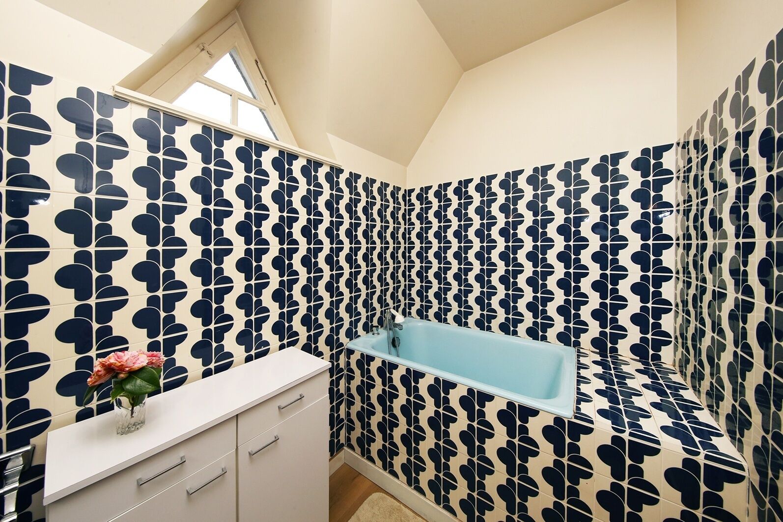 Une salle de bain avec un carrelage aux motifs géométriques, un meuble-lavabo blanc doté de tiroirs, une baignoire bleue et une lucarne au plafond.