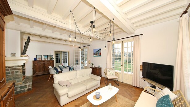 Ein großzügiges Wohnzimmer mit einem weißen Sofa, einem Couchtisch und einem Fernseher. Es gibt einen Kamin, einen Essbereich und durch große Fenster einen Blick ins Freie.