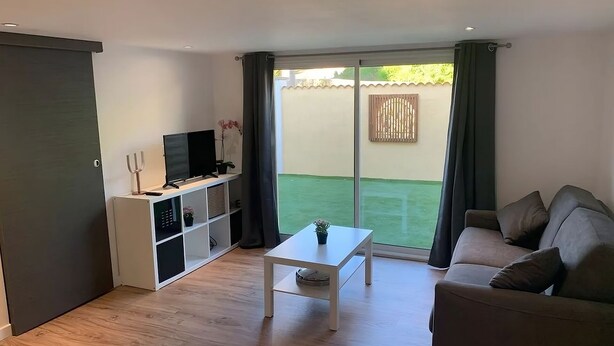 A modern living room with a sofa, a coffee table, a TV on a white stand, and a sliding glass door leading to a balcony with a green lawn. - Appartement à 5 Minutes des Plages - 42m2 - Jardin Privatif et Parking Privé (Carqueiranne)