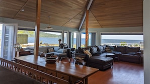 A spacious living area with a wooden ceiling, large windows offering a view of the sea, a dining table with chairs, and a comfortable seating area with sofas and cushions. - Stunning beach house with pool and hot tub. Pet friendly! Great for families!  (Bellingham)