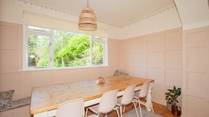 A dining area with a wooden table, four chairs, a hanging light fixture, a window with a view of greenery, and a potted plant. - Charming 3BD Oasis 3 Min Walk From Beach Front! (Dublin)