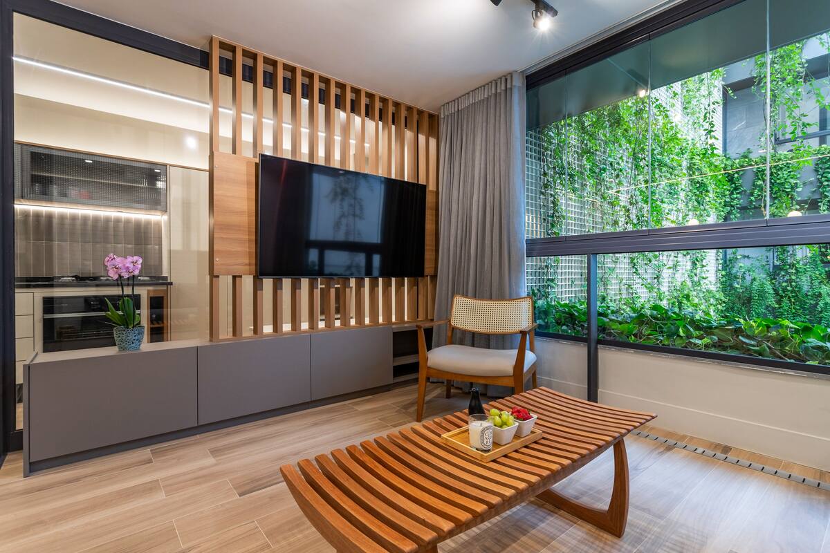 Ein modernes Wohnzimmer mit einer Holzbank, einem an der Wand montierten Fernseher und einem großen Fenster mit Blick auf Grünflächen.