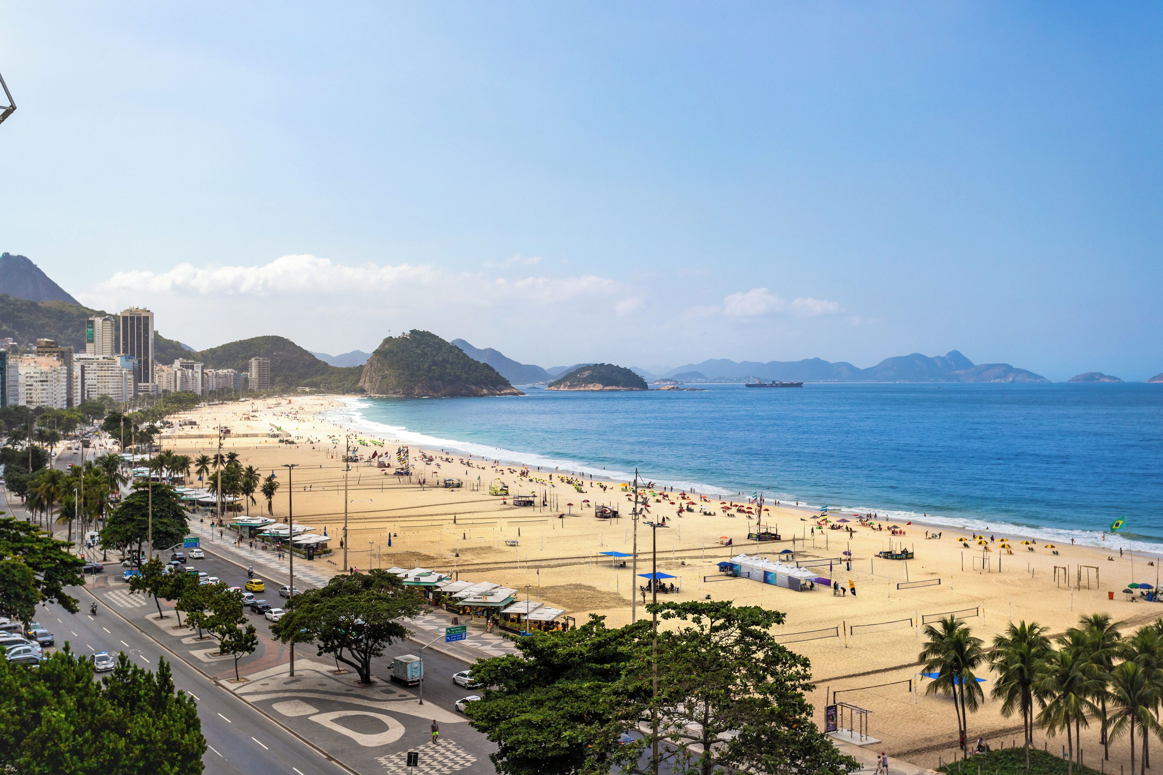 A beach with a road in the foreground, palm trees, and buildings in the background.