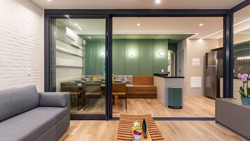 Una sala moderna con un sofá, una mesa de centro con fruta y un cuchillo, un vidrio que da a un comedor con mesa y sillas, y una cocina con refrigerador y fregadero.