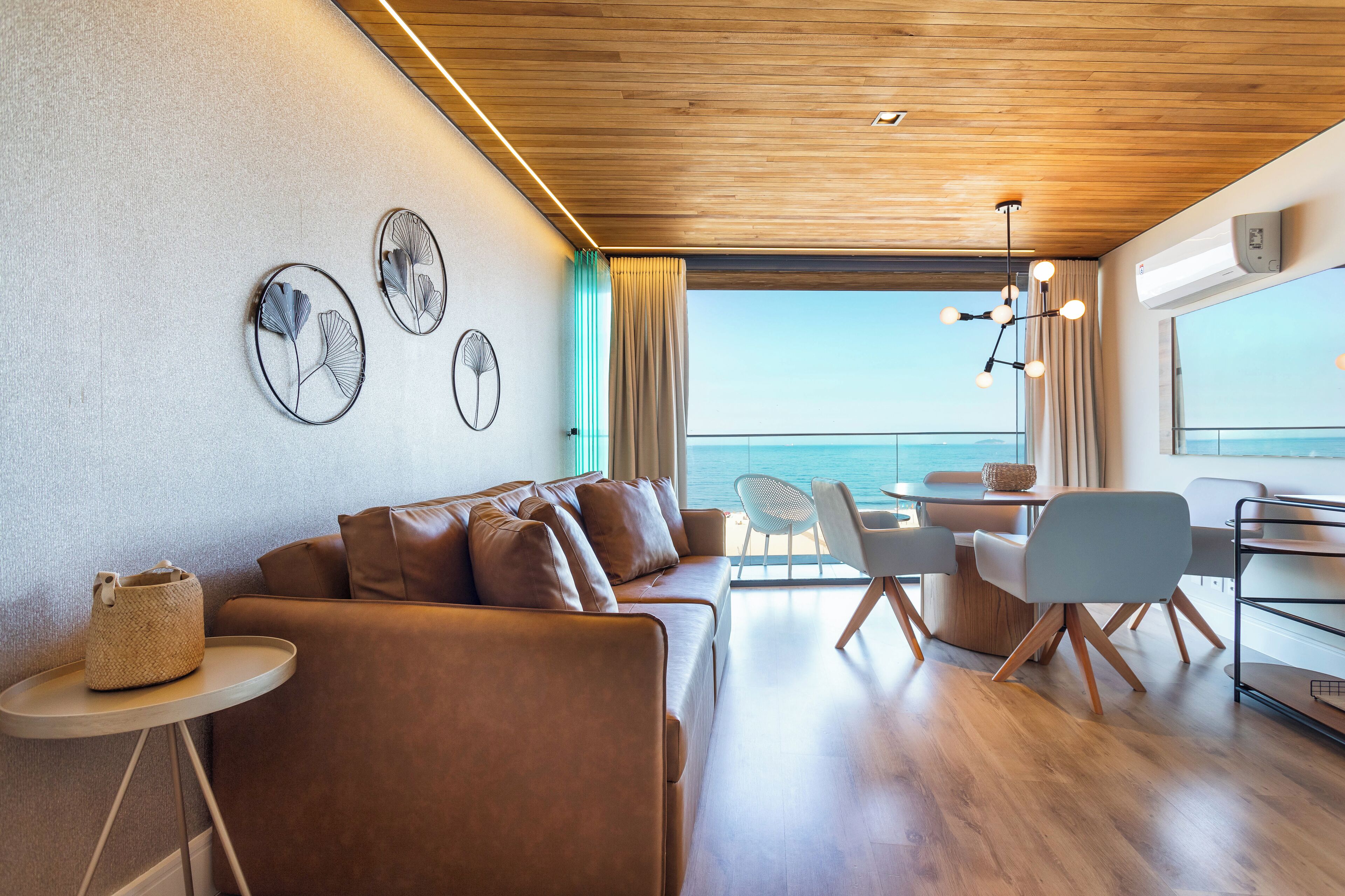 A modern living area with a wooden ceiling, a sofa, a dining table with chairs, and a view of the sea.