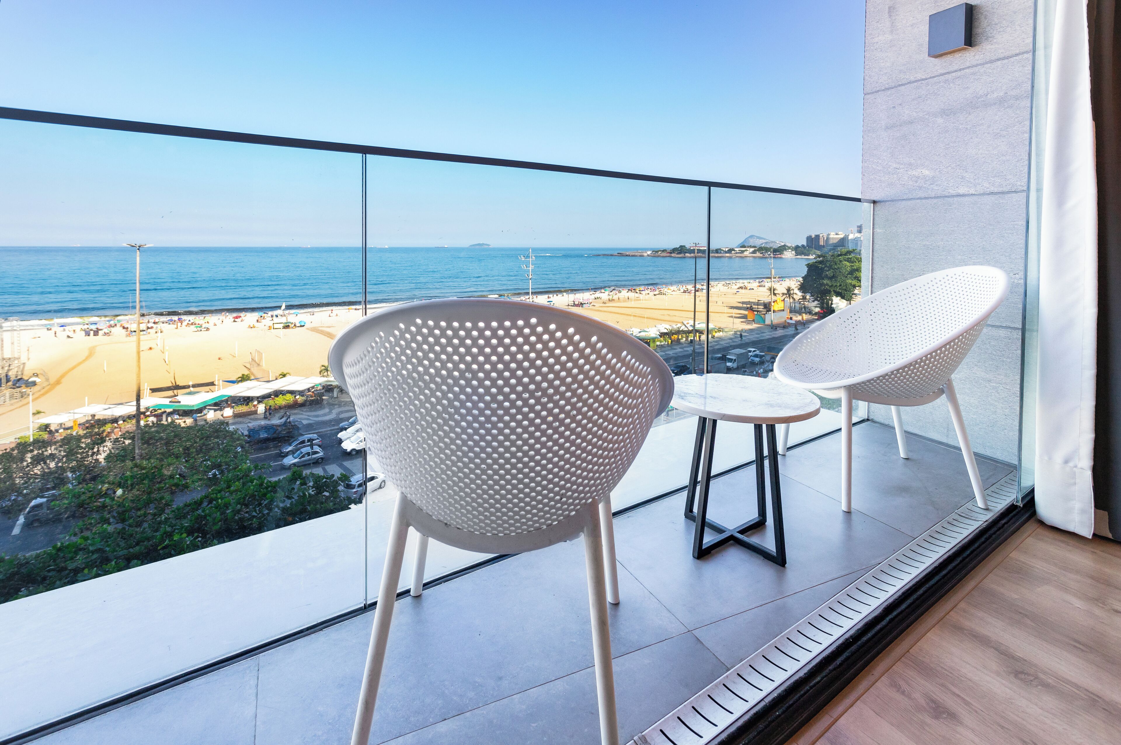 Un balcón con dos sillas blancas y una mesita pequeña, con vistas a una playa y una ciudad lejana al otro lado del mar.