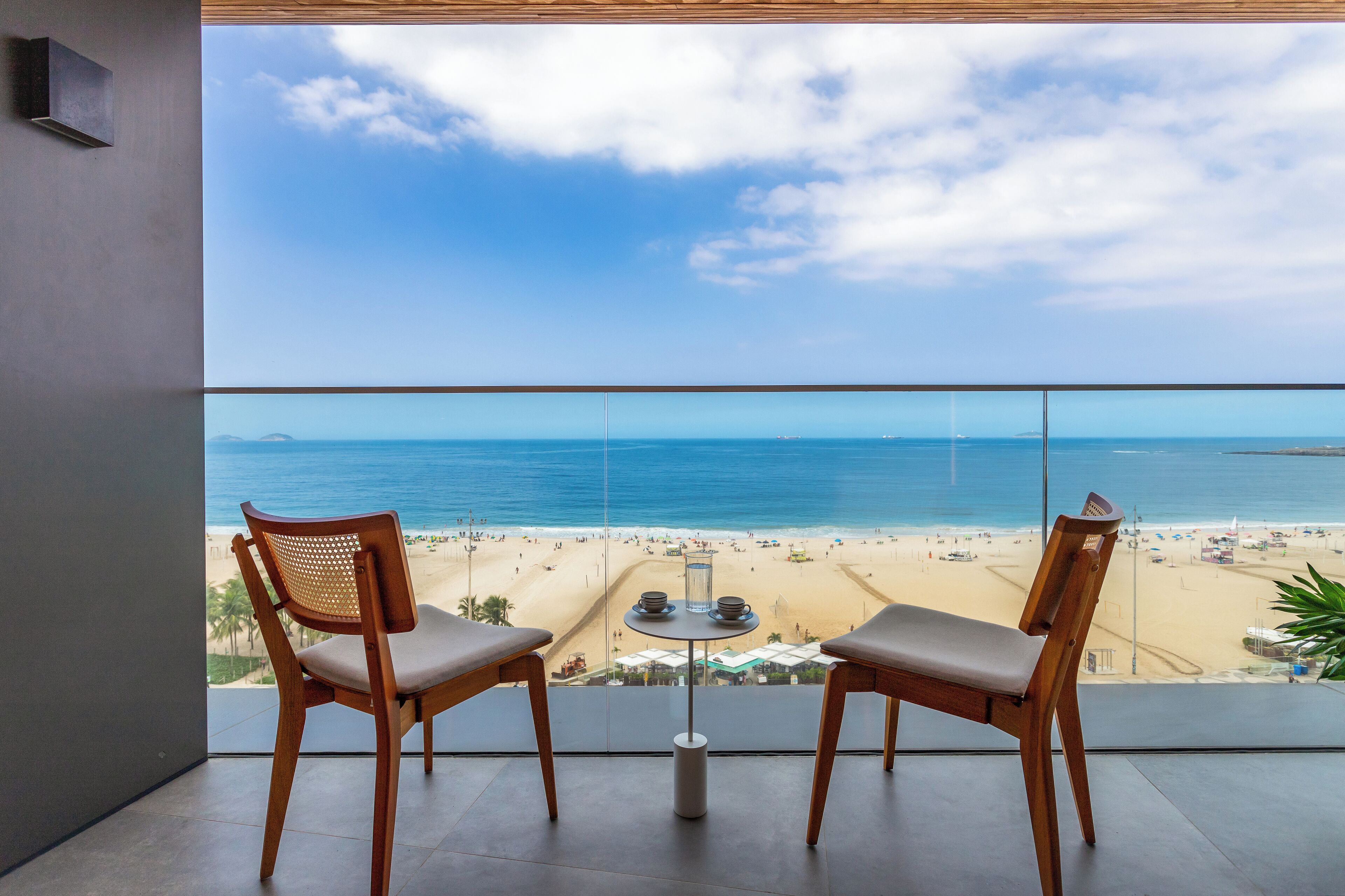 Varanda com duas cadeiras de madeira e uma mesa pequena, com vista para uma praia com pessoas e guarda-sóis.