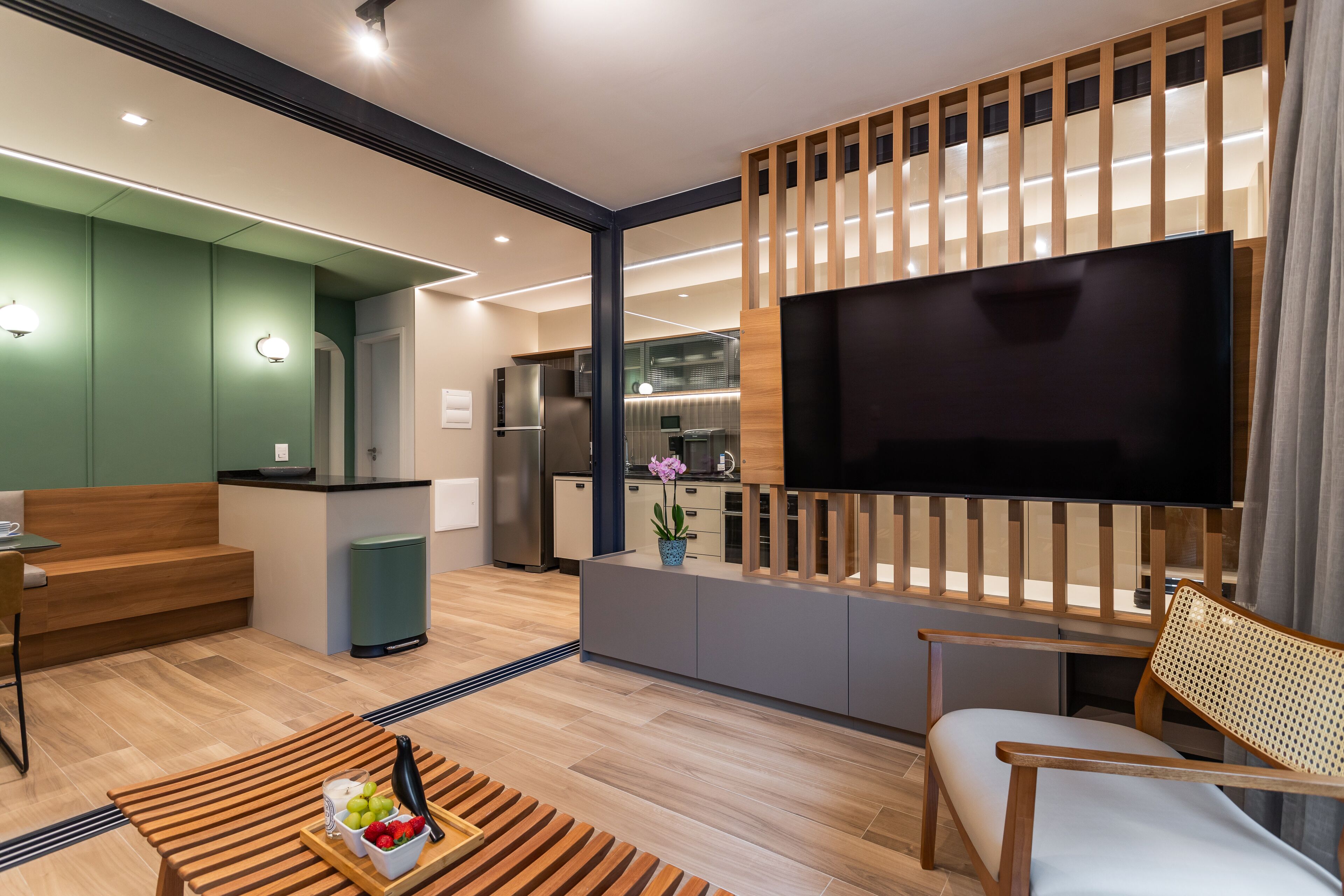 A modern living room with a flat-screen TV, wooden furniture, and a kitchen area in the background.