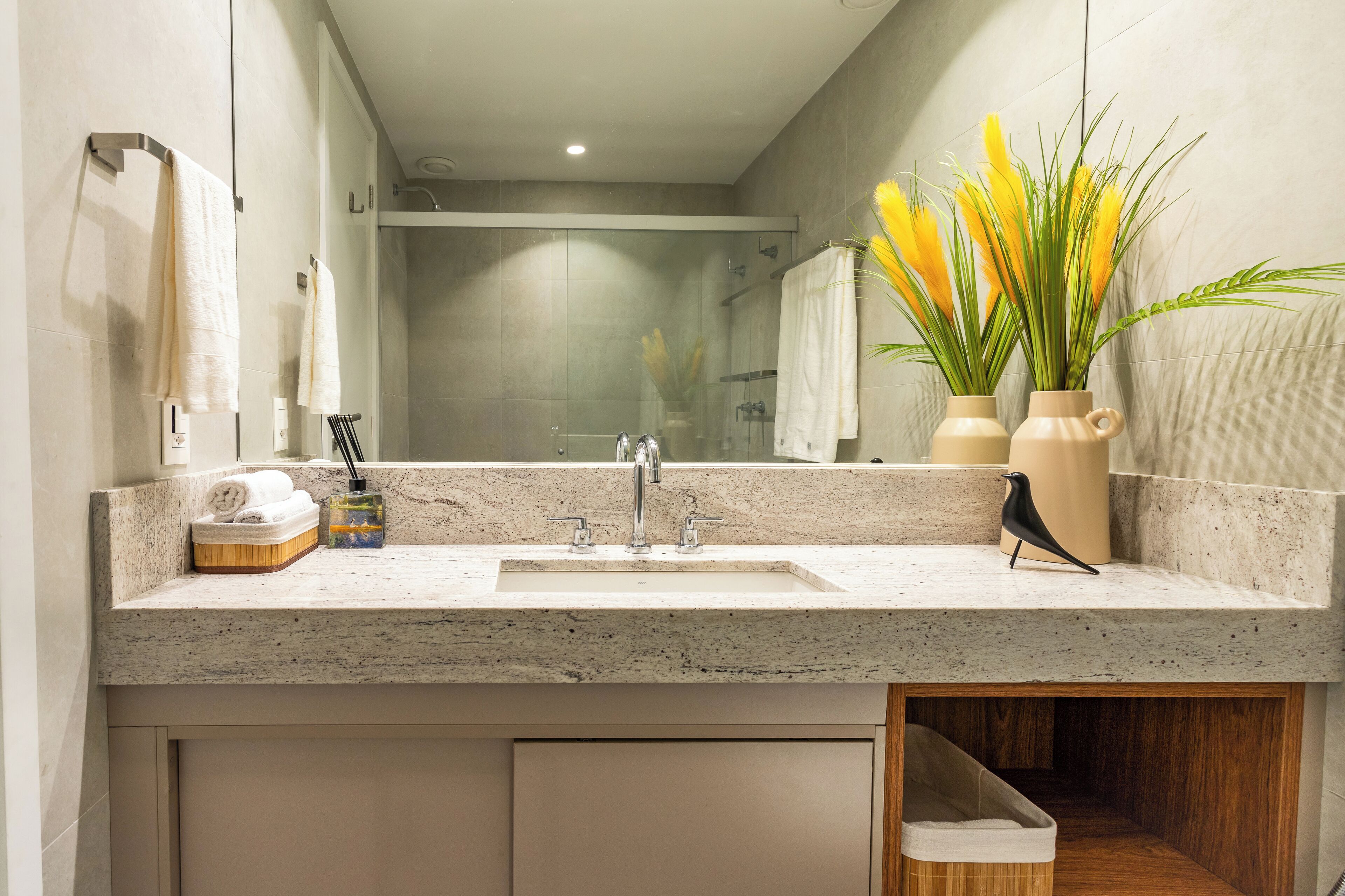 Un baño moderno con encimera de granito, lavamanos, espejo y un florero con flores amarillas.