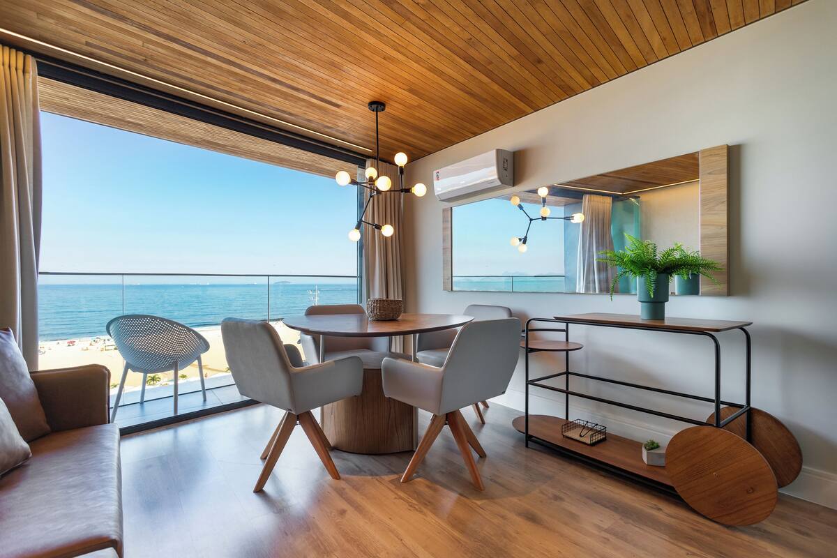 Ein modernes Wohnzimmer mit Holzbalkendecke, einem runden Esstisch mit Stühlen, einem großen Fenster mit Meerblick und minimalistischer Einrichtung.
