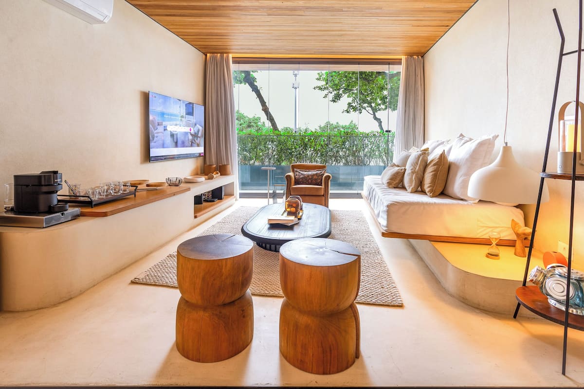 Ein modernes Wohnzimmer mit einer Couch, einem Couchtisch und Holzschemeln. Ein Fernseher ist an der Wand montiert, und es gibt ein Fenster mit Blick auf Grünflächen.