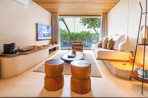 A modern living room with a sofa, coffee table, and wooden stools. A television is mounted on the wall, and there is a window with a view of greenery. - Unhotel (Rio de Janeiro)