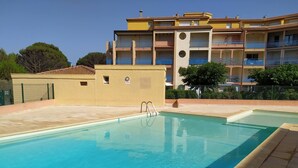 A swimming pool with a clear blue water, surrounded by a tiled deck. In the background, there is a multi-story building with balconies and a yellow facade. - V0742 - Apt 4 pers Vias Plage parking piscine collective (Vias)