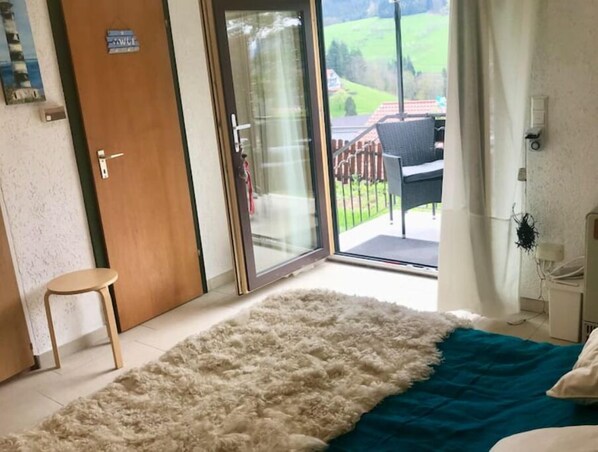 A room with a large fluffy rug, a wooden door, a glass door leading to an outdoor area with a view of a green landscape, and a small stool.