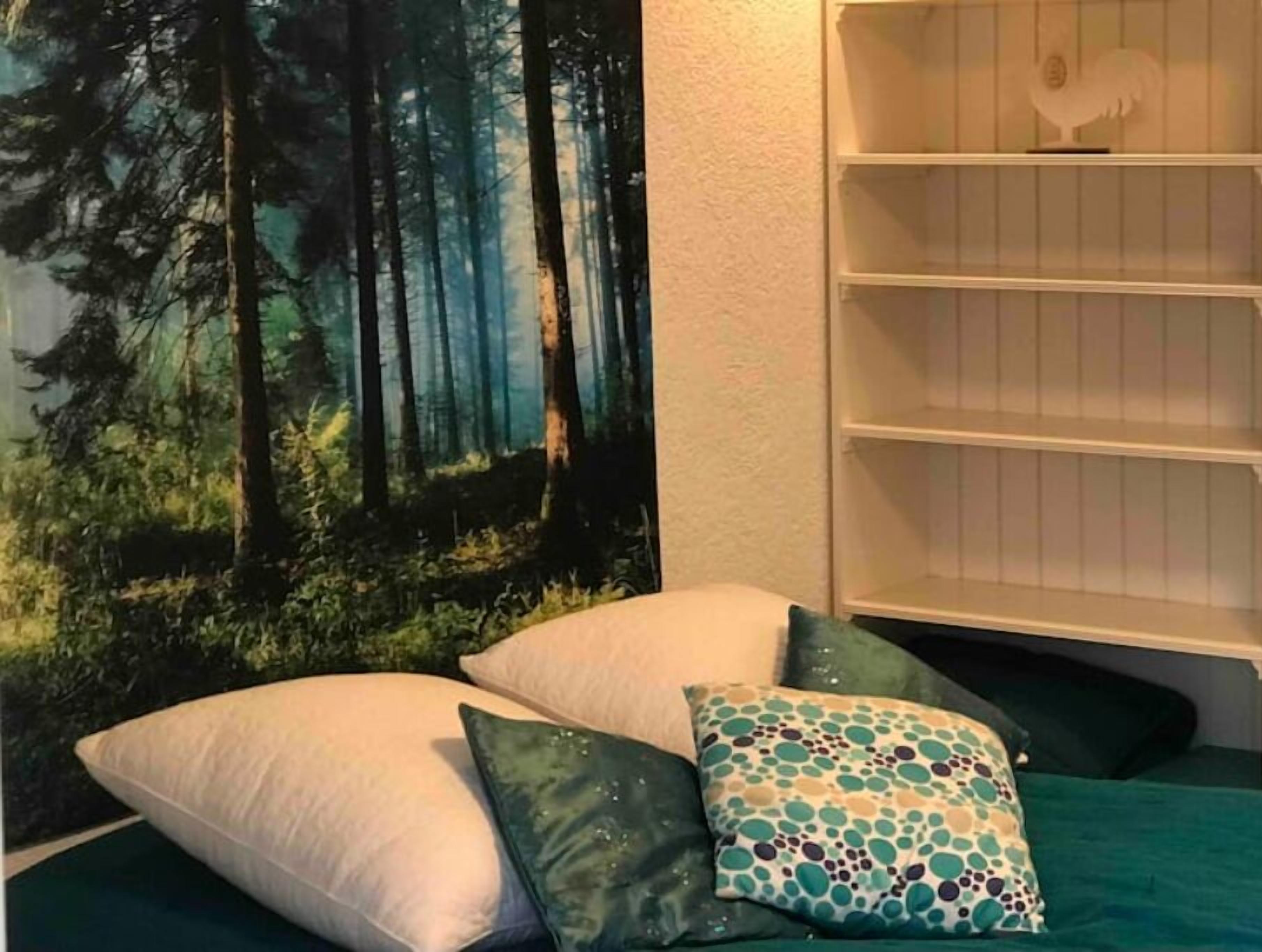 A bedroom with a forest-themed tapestry, a bookshelf, and a bed with green bedding and patterned pillows.