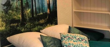 A bedroom with a forest-themed tapestry, a bookshelf, and a bed with green bedding and patterned pillows.