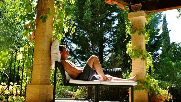 A man relaxing on a sun lounger under a pergola with greenery around.