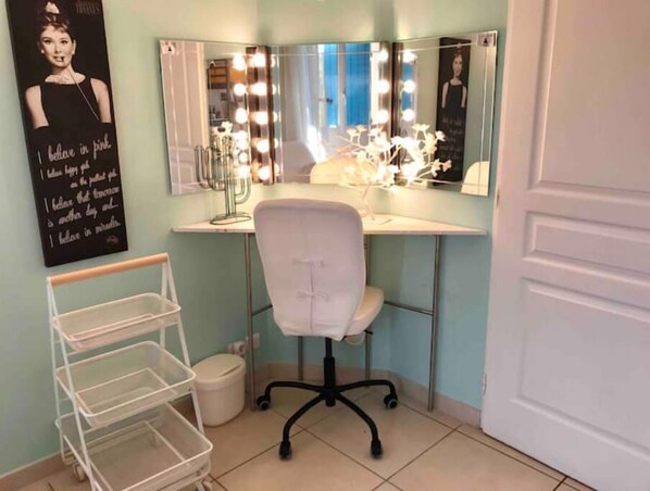 A room with a vanity, mirror, and lights. There is a chair and a white shelf unit.