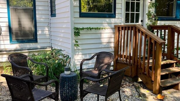 Une maison avec une véranda, trois chaises en osier, une petite table et un escalier en bois.