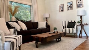 Un salon avec un canapé marron foncé, des fauteuils à rayures, une table basse en bois et une lampe. Des tableaux encadrés ornent le mur, et une fenêtre est agrémentée de rideaux.