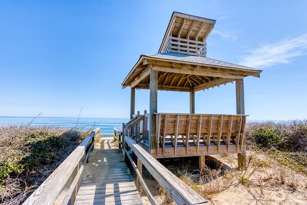 A wooden beach boardwalk leading to a wooden beach shelter with a slanted roof. - Oceanfront condo with great views & community pool! (Duck)