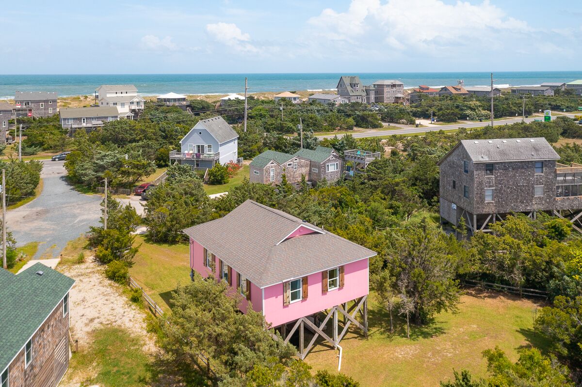 A coastal residential area with houses, including a pink house on stilts, surrounded by greenery and a beach in the background.