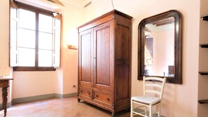 Ein Zimmer mit großem Holzschrank, einem weißen Stuhl, einem großen Spiegel und einem Fenster mit geöffneten Rollläden.