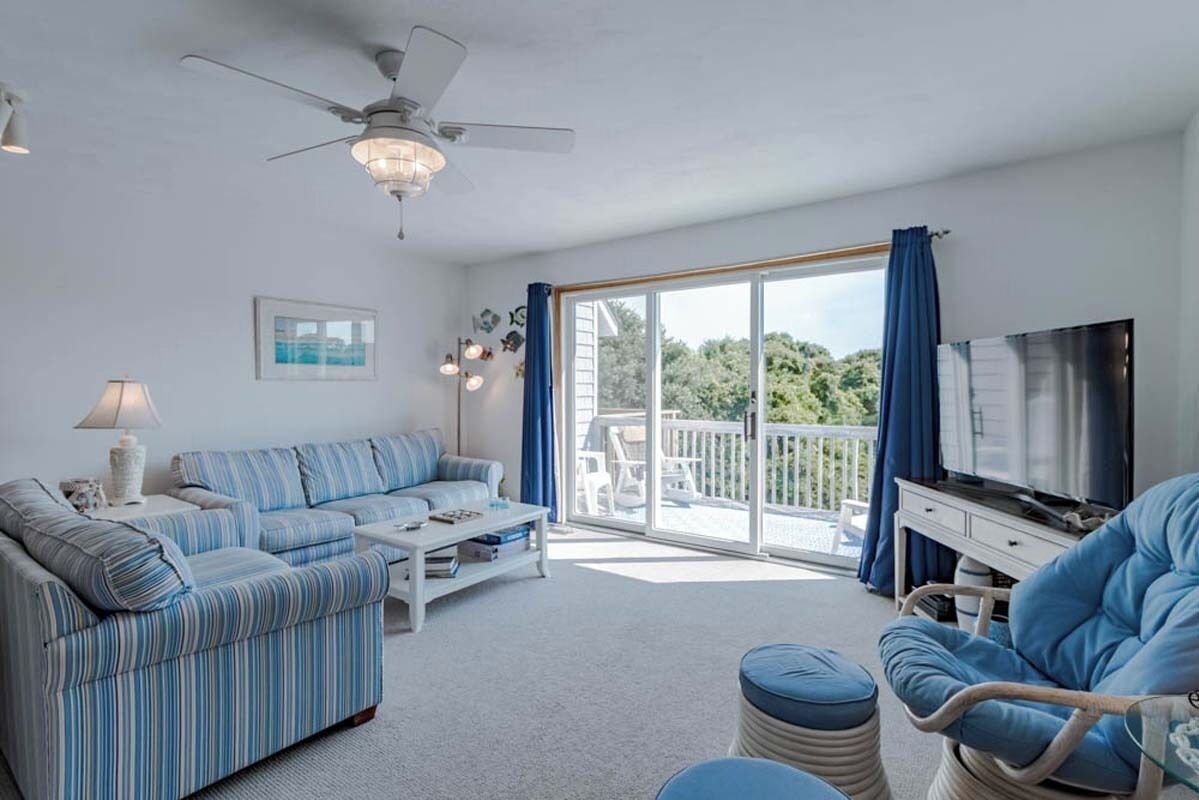 A living room with a sofa, coffee table, and a television. There is a balcony with a view of greenery.