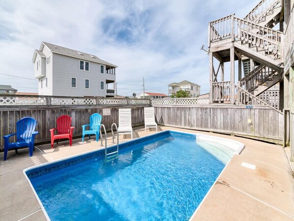 Una piscina rectangular con cubierta de madera, rodeada por una valla de madera. Hay cuatro sillas Adirondack en colores azul, rojo y blanco.