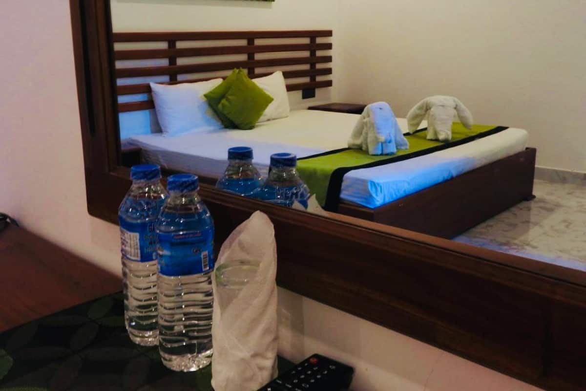 A hotel room with a bed, pillows, a blanket, a remote control, and several water bottles.