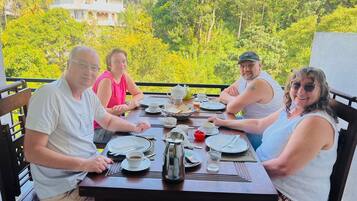 A group of people seated at a table with plates, cups, and a teapot, overlooking a lush green landscape.