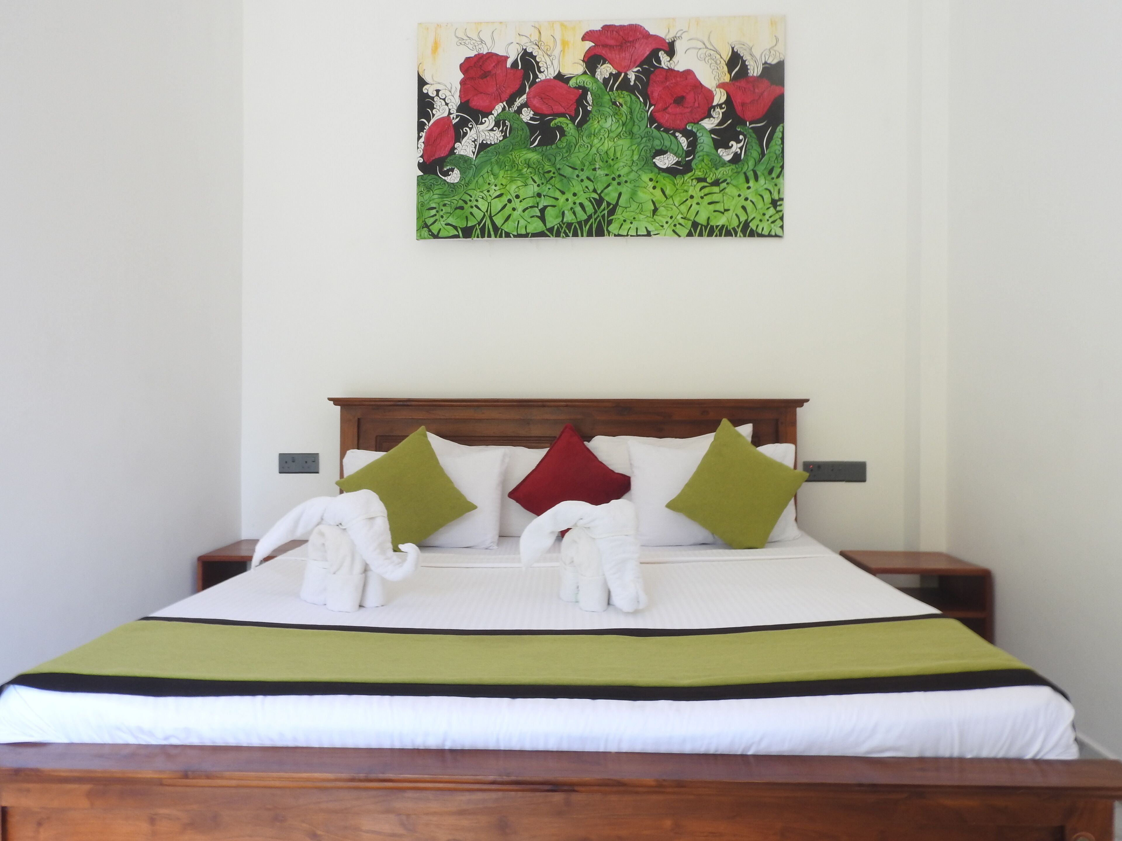 Een net opgemaakt bed met een groen-witte sprei, een houten hoofdbord, en een schilderij van rode bloemen aan de muur.