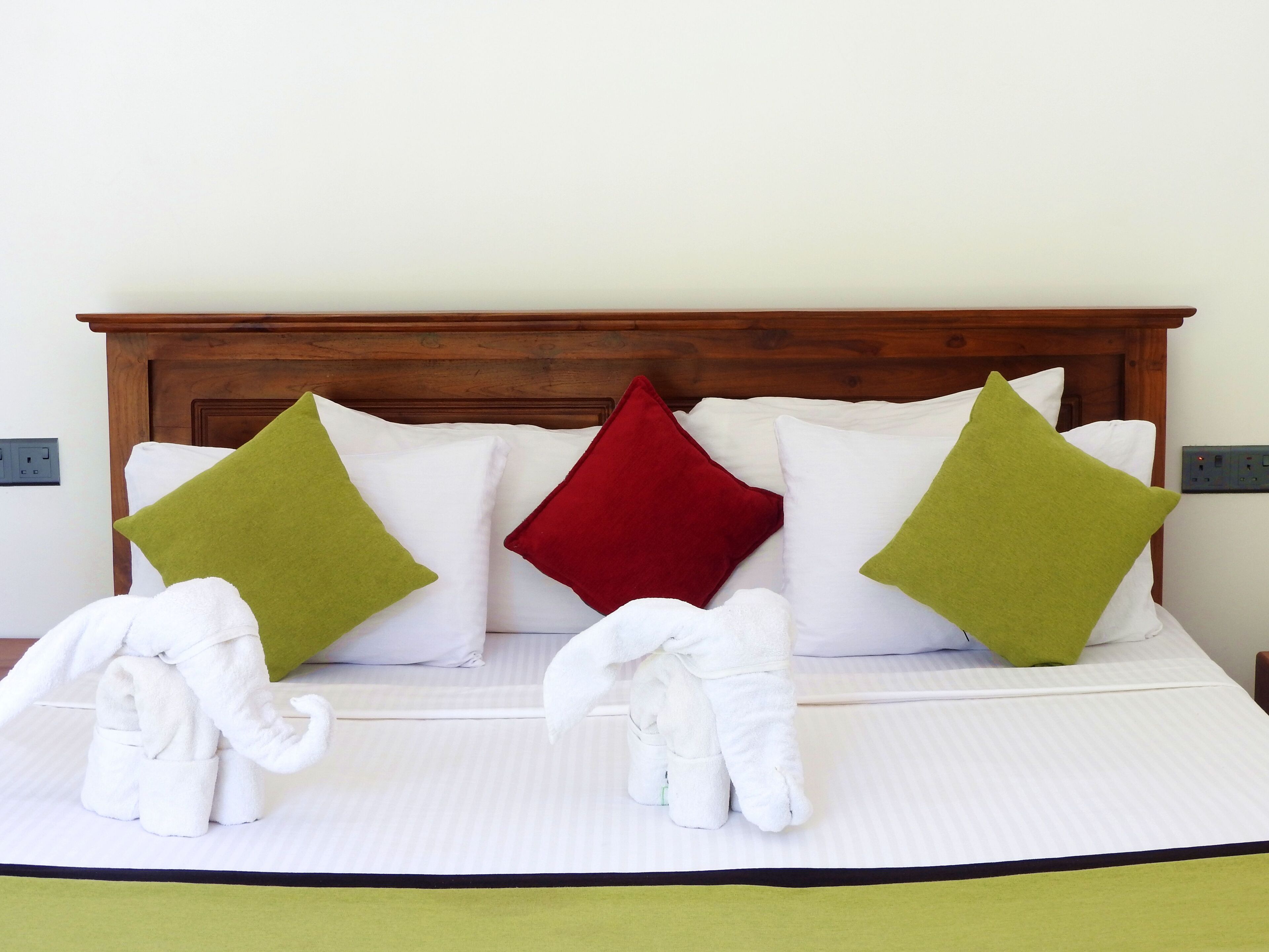 A neatly made bed with white linens, green and red decorative pillows, and two white elephant-shaped bathrobes.