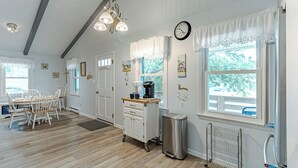 A room with a wooden floor, white walls, a dining table with chairs, a kitchen area with a coffee maker, a trash bin, and a window with a view of trees.