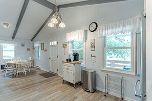 Una habitación con piso de madera, paredes blancas, una mesa de comedor con sillas, un área de cocina con cafetera y una ventana con vista a árboles.