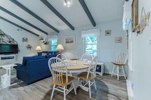 A dining area with a round wooden table, white chairs, a blue sofa, and a television in the background. - Pet-friendly oceanside home in Nags Head by Sun Realty (Nags Head)