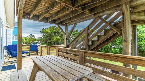 A wooden deck with picnic tables and blue chairs, a staircase, and a view of trees and a building.