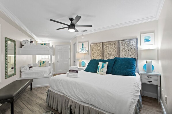 A bedroom with a large bed, a ceiling fan, a bench, and a door. - Amalfi Coast 209A - Gulf View, Swimming Pool, Tennis Court, Near Shops + Dining! (Miramar Beach)