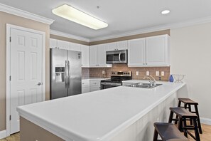 A modern kitchen with white cabinetry, stainless steel appliances, a large island with stools, and a tiled backsplash. - Amalfi Coast 209A - Gulf View, Swimming Pool, Tennis Court, Near Shops + Dining! (Miramar Beach)