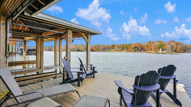 A wooden deck with Adirondack chairs overlooking a lake, surrounded by autumn trees. - Hot Tub, Boat Dock, Large Deck, Firepit, Tiki bar, Pool! (Littleton)