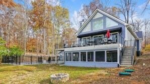 A two-story house with a balcony, a stone fire pit, and a view of autumn trees. - Hot Tub, Boat Dock, Large Deck, Firepit, Tiki bar, Pool! (Littleton)