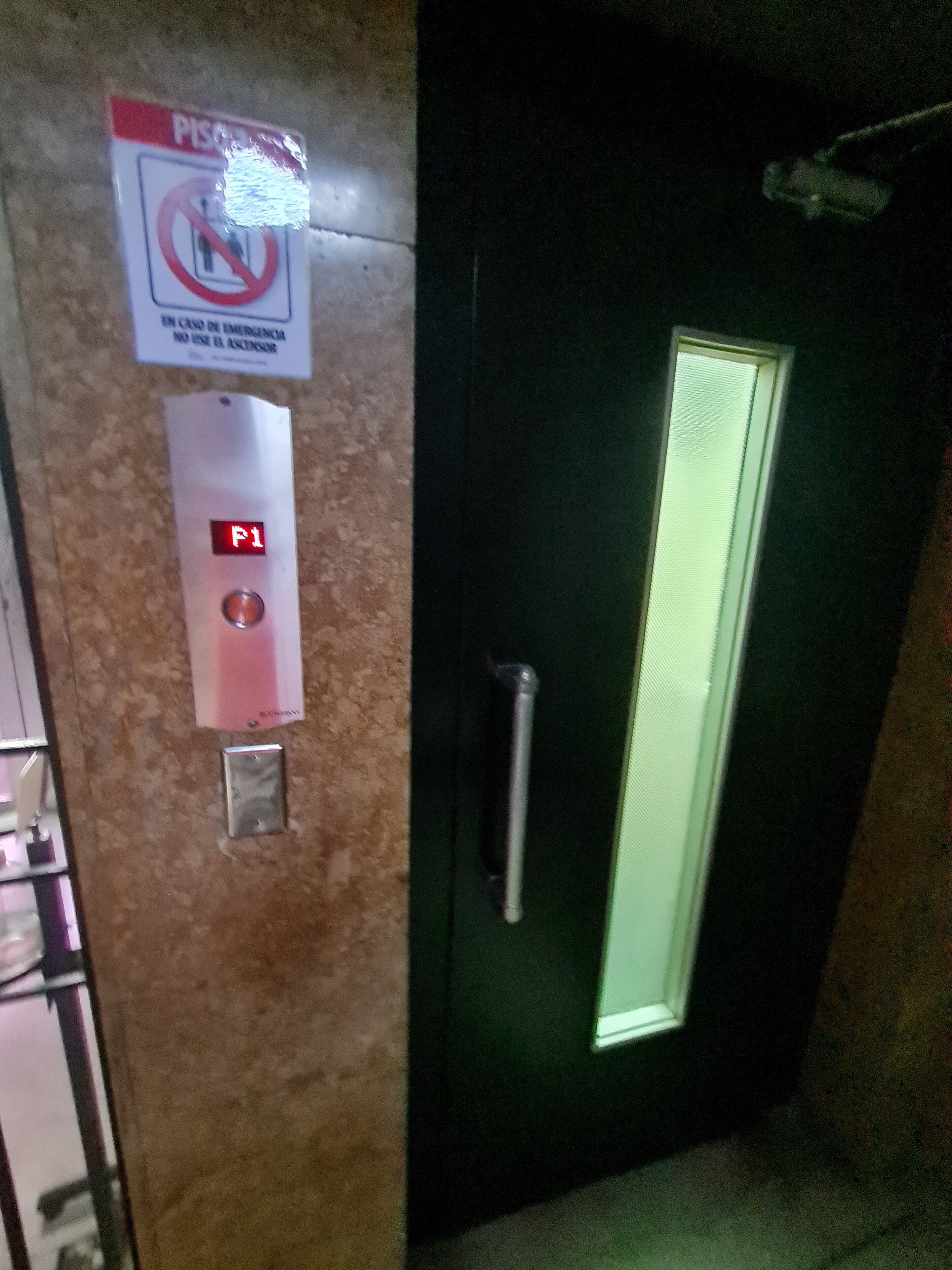 Een deur met een verticaal raam, een rode knop voor noodgevallen en een bord waarop staat dat de lift niet gebruikt mag worden bij noodsituaties.