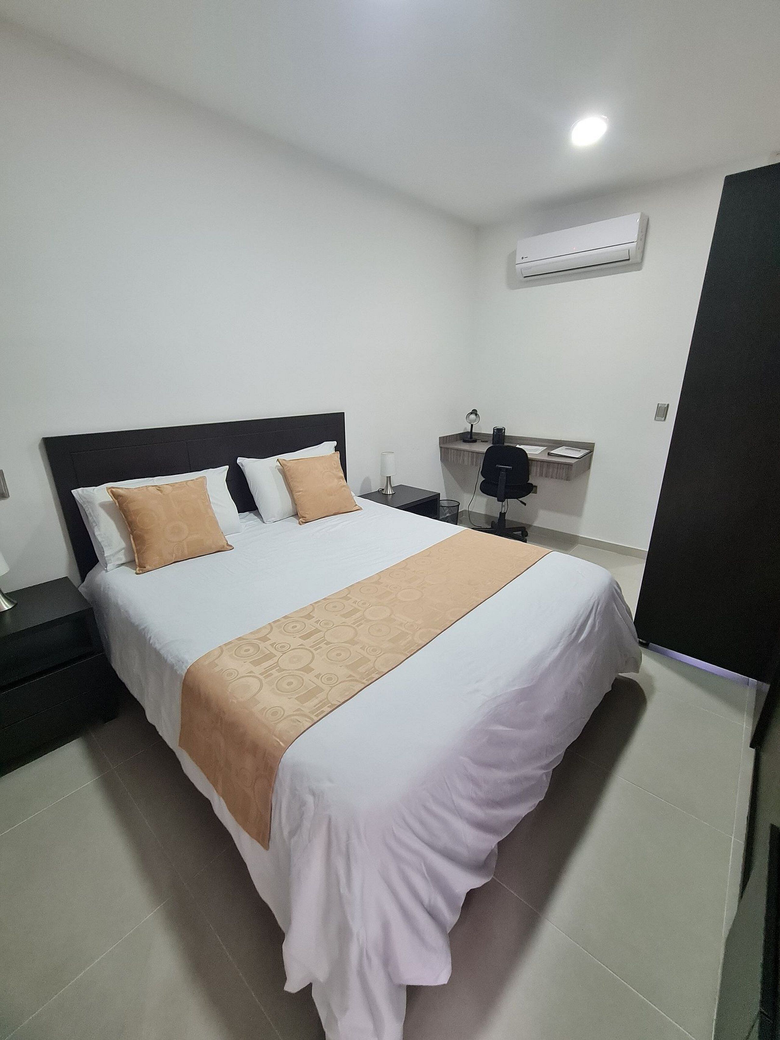 A neatly made bed with a white and beige bedspread, a black headboard, and two pillows. There is a bedside table with a lamp and a small mirror. An air conditioning unit is mounted on the wall above the bed.