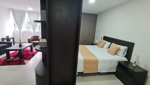 Ein modernes Hotelzimmer mit einem großen Bett, einer dunklen Holzablage, einer kleinen Sitzecke mit Stuhl und Tisch sowie einem Fenster mit Vorhängen.