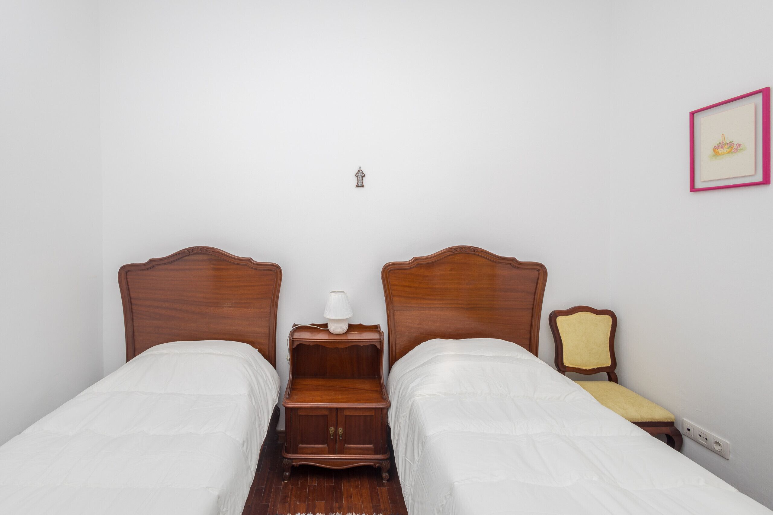 A bedroom with two single beds, a nightstand with a lamp, a chair, and a wall-mounted light switch.