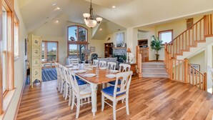 A spacious dining area with a wooden table set for six, surrounded by white chairs with blue cushions. A chandelier hangs from the ceiling, and a staircase with wooden railings leads to an upper level. - Pet-friendly canalfront home in Avon w/ private pool and hot tub! (Avon)